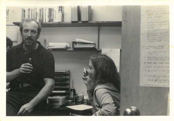 A man sits on a desk and drinks a beer. A woman, seated, smokes a cigarette.