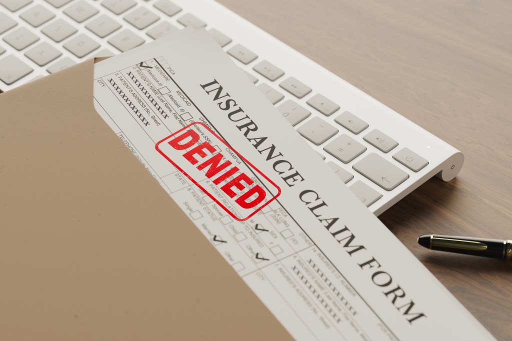 A denied health insurance claim form on a wooden table next to a keyboard and pen.