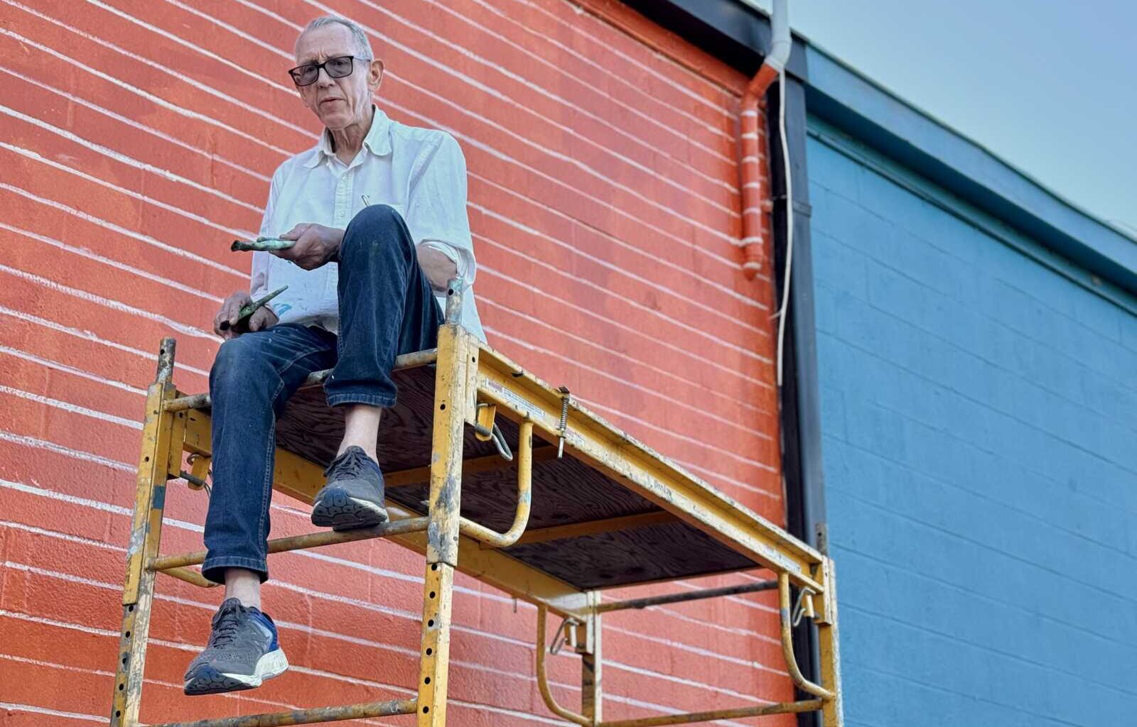 Older man wearing glasses sits on yellow scaffold painting a red brick wall with a paintbrush and roller while blue siding is visible nearby.