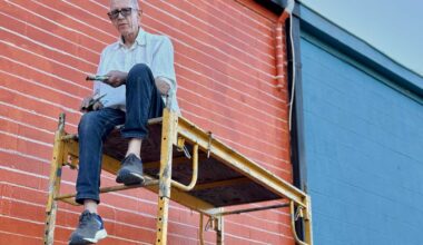 Older man wearing glasses sits on yellow scaffold painting a red brick wall with a paintbrush and roller while blue siding is visible nearby.