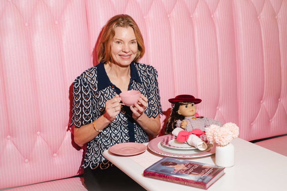Author Fiona Davis, 59, sits with her book and her Samantha doll at the American Girl café. She has chin-length blond hair and is wearing a woven navy blouse while sitting in a pink booth and holding a pink teacup.
