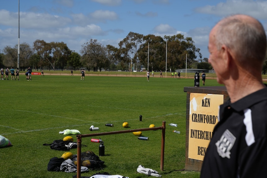 A man in a black polo shirt standing on the side lines of an oval, watching football players train