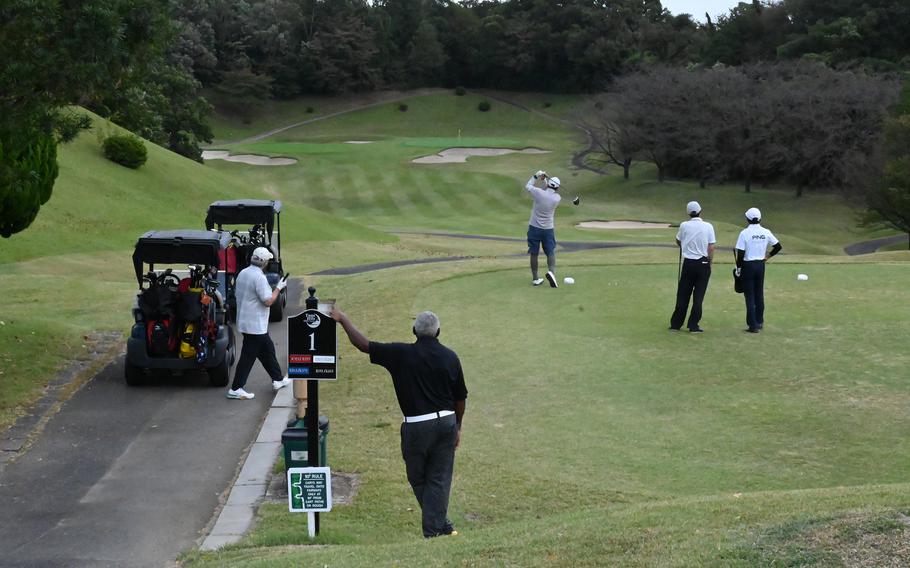 Golfers gather at the first tee of a tree-lined golf course, with one player driving toward the fairway while others watch near golf carts and the tee marker.
