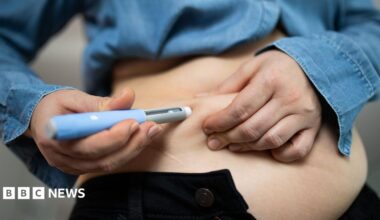 A person injecting a medication into their stomach - they are wearing a denim shirt and black trousers