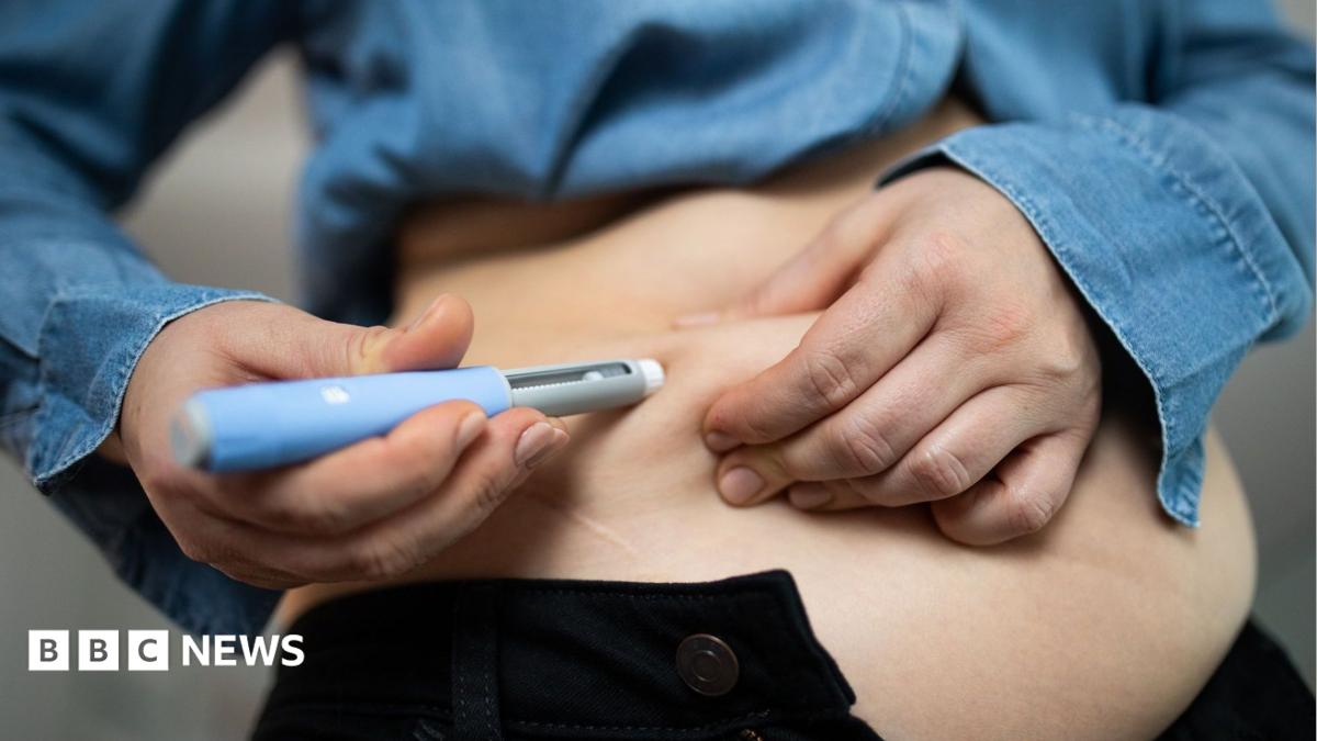 A person injecting a medication into their stomach - they are wearing a denim shirt and black trousers