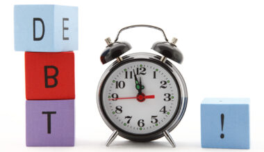 A photo of an alarm clock with multicolored building blocks on either side of it. The three blocks on the left side spell out "D" "B" "T" and the one on the right has an exclamation point on it.