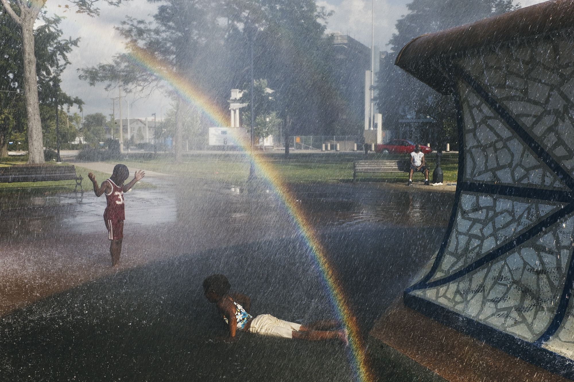 A rainbow arcs through the spray of a park sprinkler, with a child raising their hands on the left, another child lying on the wet ground and a man sitting on a bench in the background.