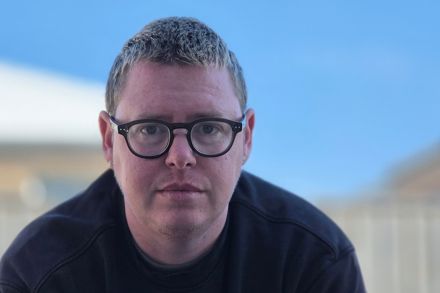 A person wearing a black shirt and black glasses looks seriously at the camera.
