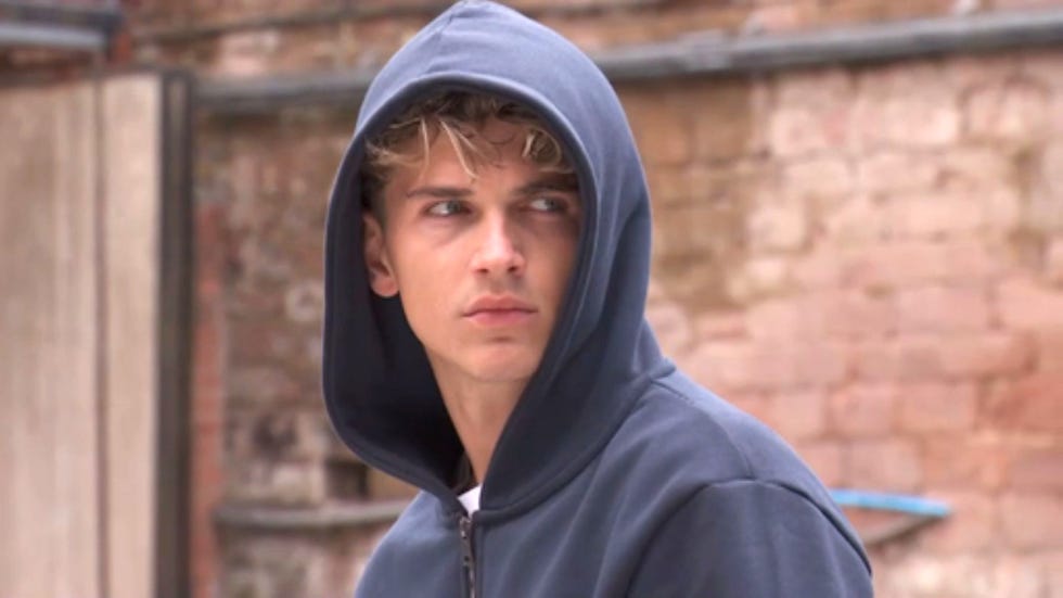 hollyoaks lucas hay person in a blue hoodie looking to the side against a brick wall background.