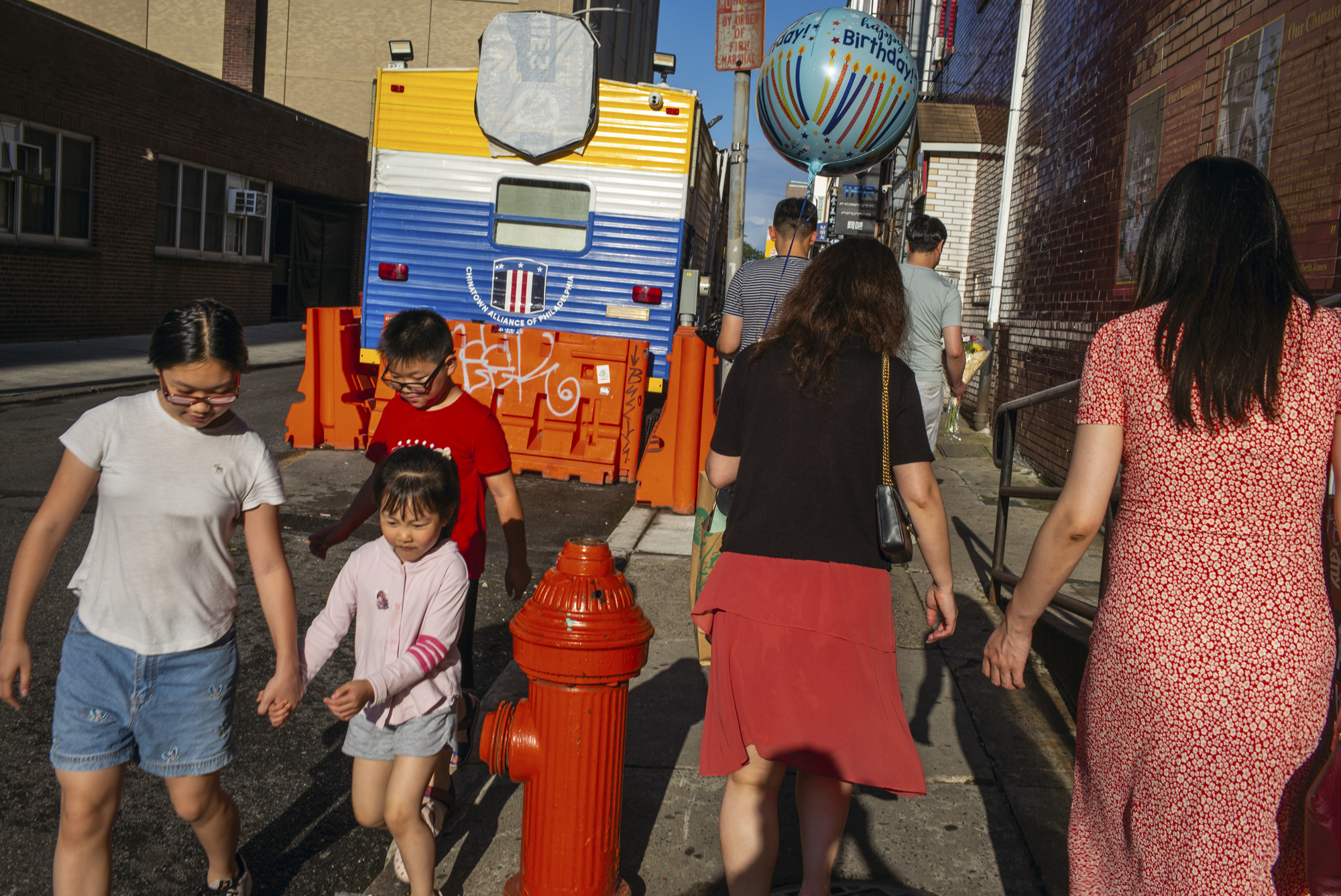 Children and adults walk along a sunlit Chinatown street past a bright red fire hydrant, with a girl leading a smaller child by the hand, women seen from behind in red and black