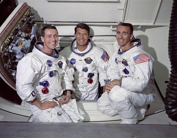 A color photograph shows three smiling men wearing white spacesuits adorned with patches and American flag emblems, posing closely together inside a white spacecraft interior. The suit details, including blue and red metallic connectors, stand out brightly under clear, even indoor lighting. An open hatch door with metallic mechanical components frames the left side of the background.