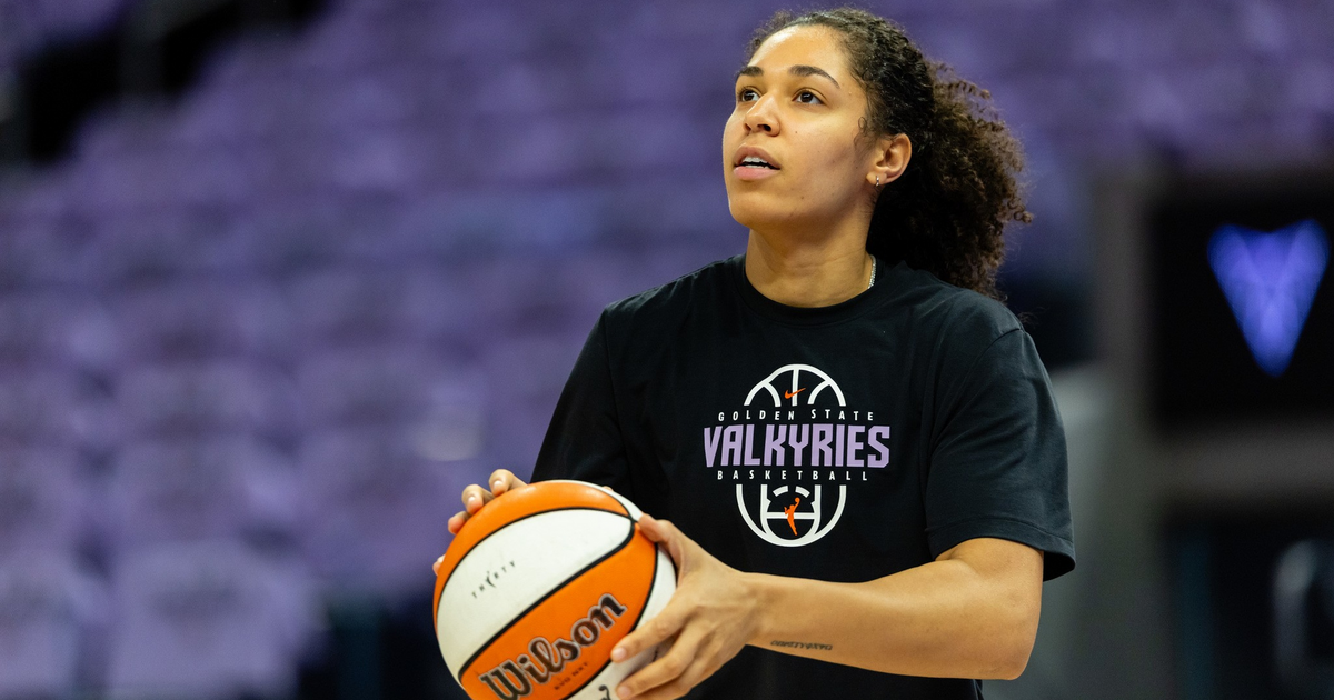A woman wearing a black Golden State Valkyries basketball shirt holds a Wilson basketball, preparing to shoot, with an empty purple-seated background.
