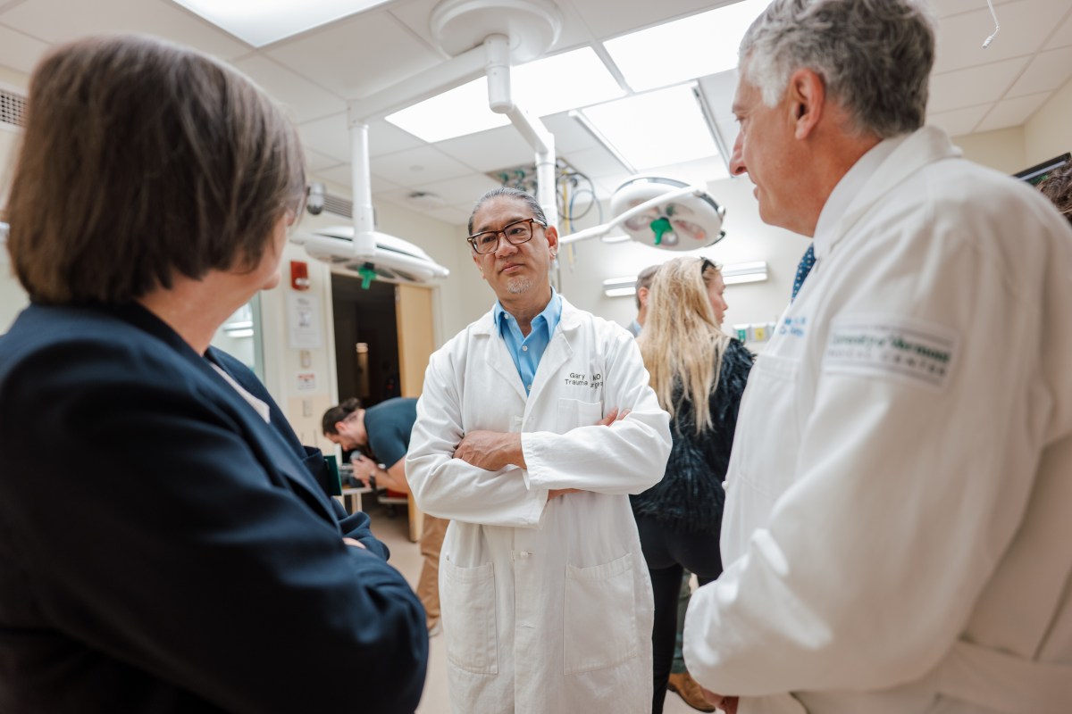 Three medical professionals, two in white lab coats and one in a dark jacket, stand talking in a hospital room with medical equipment and bright lighting.