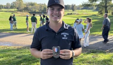 Smiling young golfer in a dark polo and cap holds a medal on a sunny golf course in the foreground.