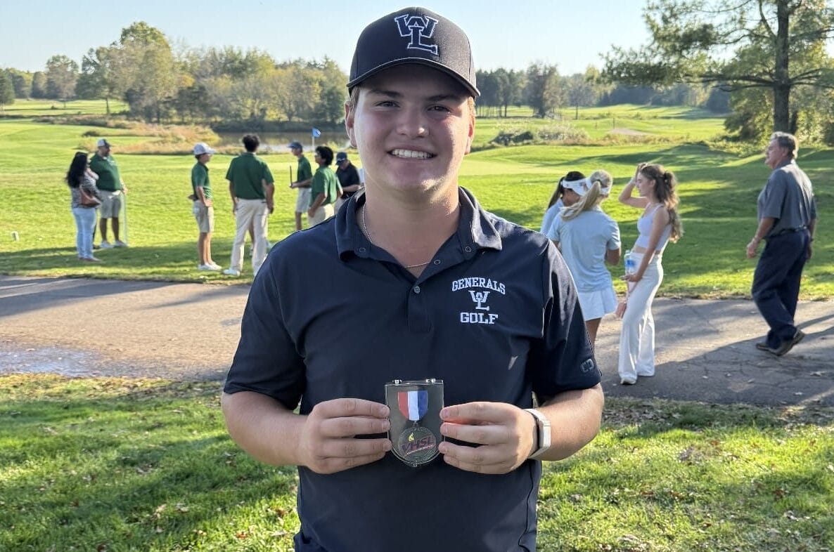 Smiling young golfer in a dark polo and cap holds a medal on a sunny golf course in the foreground.