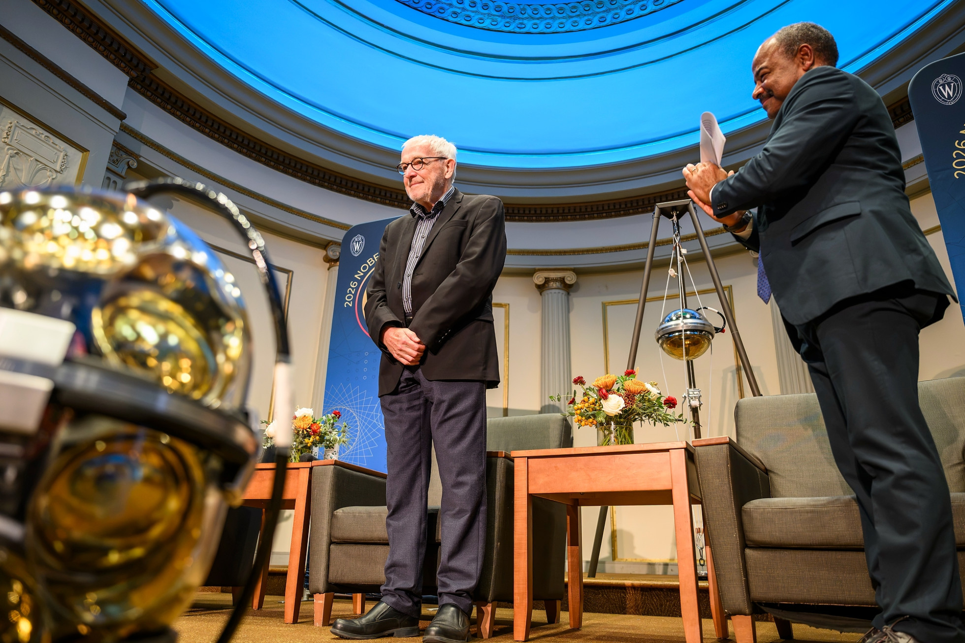 Nobel Laureate Francis Halzen smiling on a stage with blue light backdrop UW–Madison Interim Chancellor Wilcots clapping.