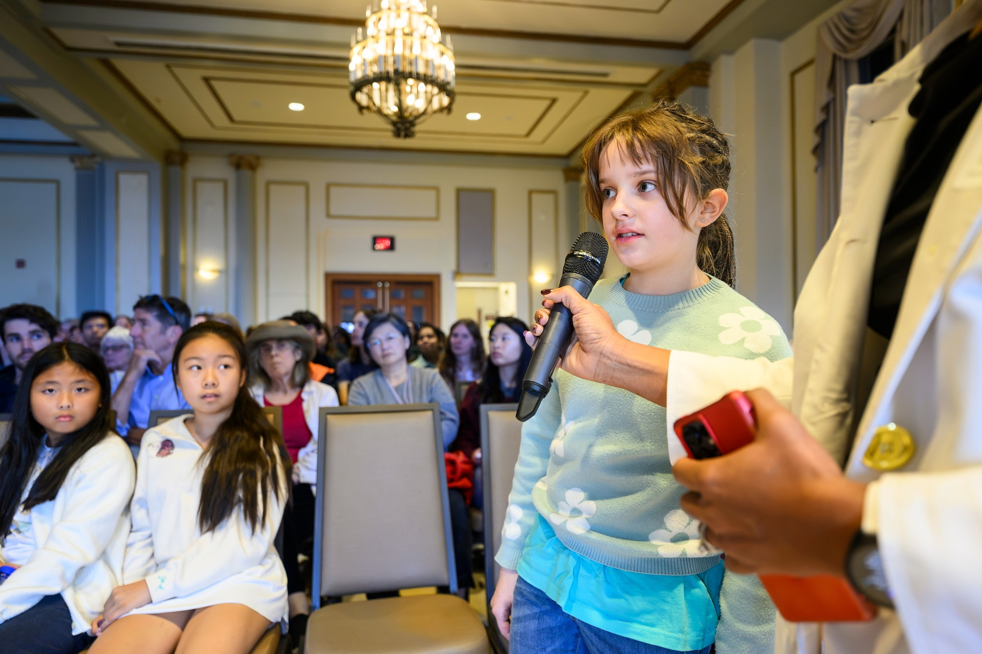 A child speaking into a microphone with a crowd of onlookers behind them