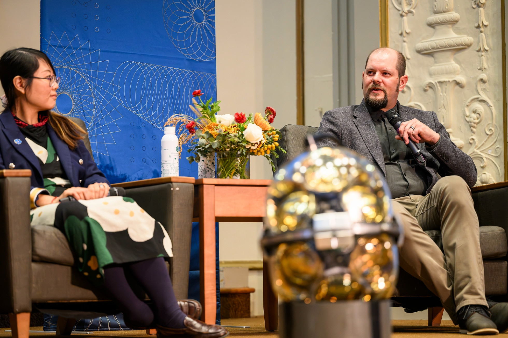 Two panelists on a stage with flowers and a blue banner in the background