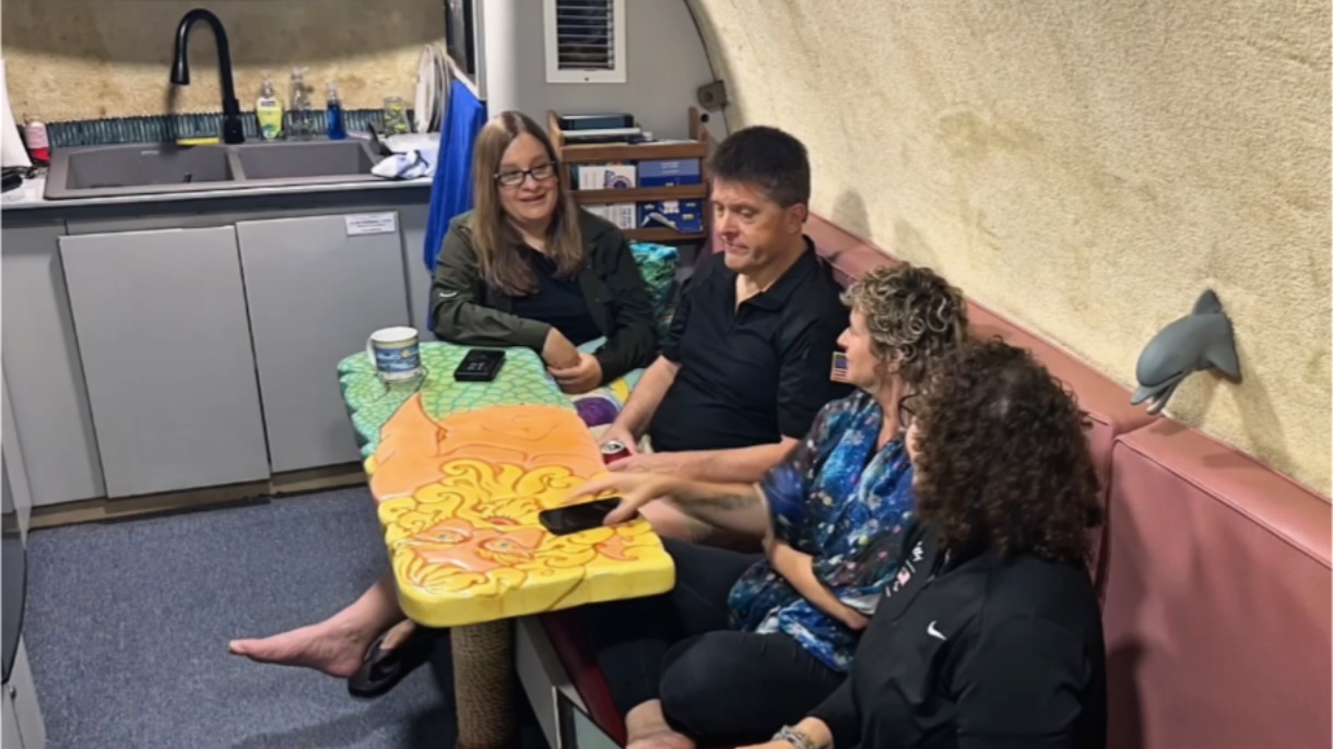 three women and one man sit on a red bench in a small kitchen with blue carpet