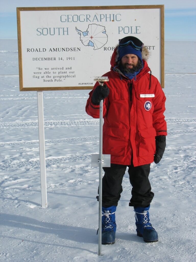 bearded man in red coat standing on snow in front of sign reading 'Geographic South Pole'