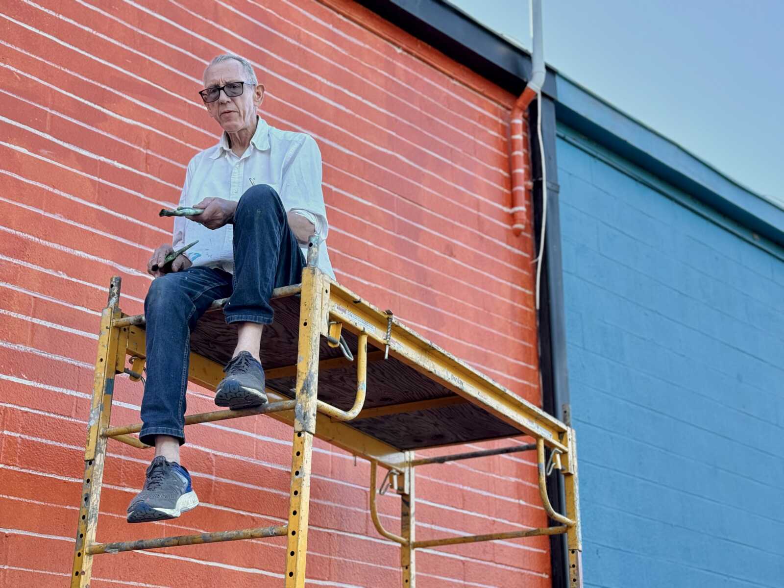 Older man wearing glasses sits on yellow scaffold painting a red brick wall with a paintbrush and roller while blue siding is visible nearby.