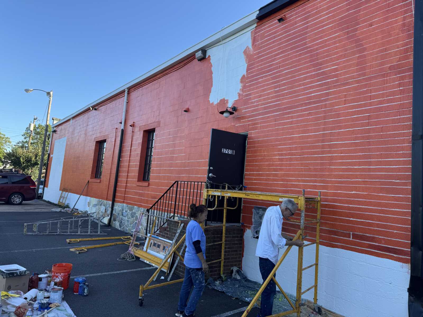 Two workers repainting the exterior of an orange brick building; a blue patch is being painted above a doorway, with paint cans and scaffolding on the ground nearby