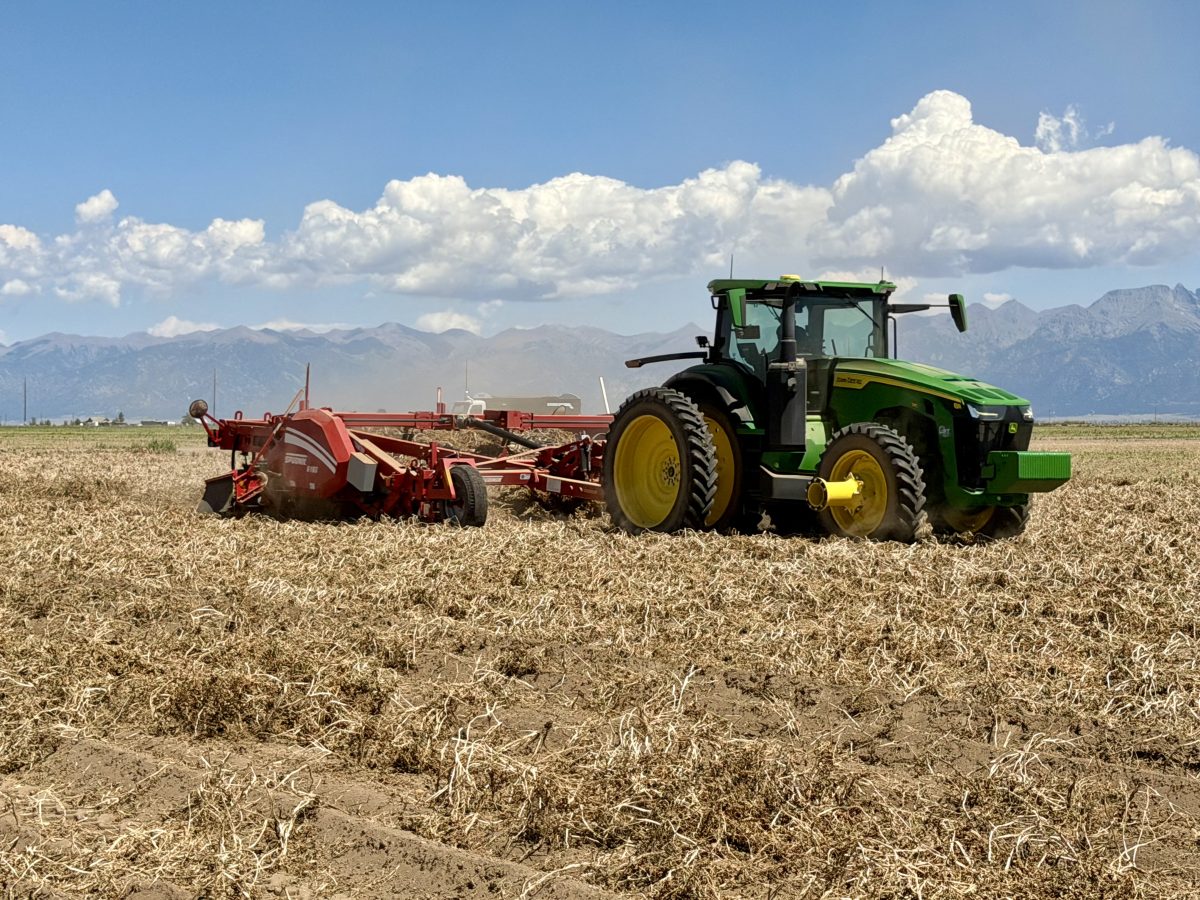 A green tractor pulls red farming equipment across a dry, harvested field with mountains and clouds in the background.