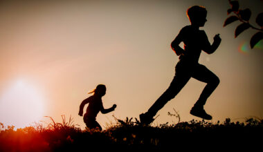 Silhouettes of two children running across a grassy hilltop at sunset.