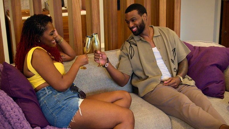 "Two people sit on a couch in a cozy living room, smiling and toasting with gold glasses. They relax among purple pillows, suggesting a casual, friendly atmosphere."