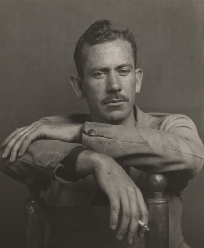 A black-and-white portrait of a man with a mustache, leaning his arms on the back of a chair while holding a cigarette.