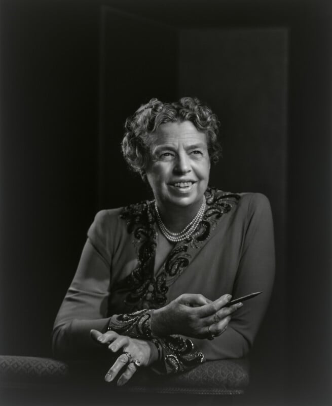 A black-and-white portrait of Eleanor Roosevelt, wearing a beaded dress and pearls, holding a pencil while looking to the side.