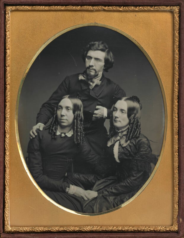 A daguerreotype portrait of a bearded man standing behind two seated women, all dressed in mid-19th-century formal attire.