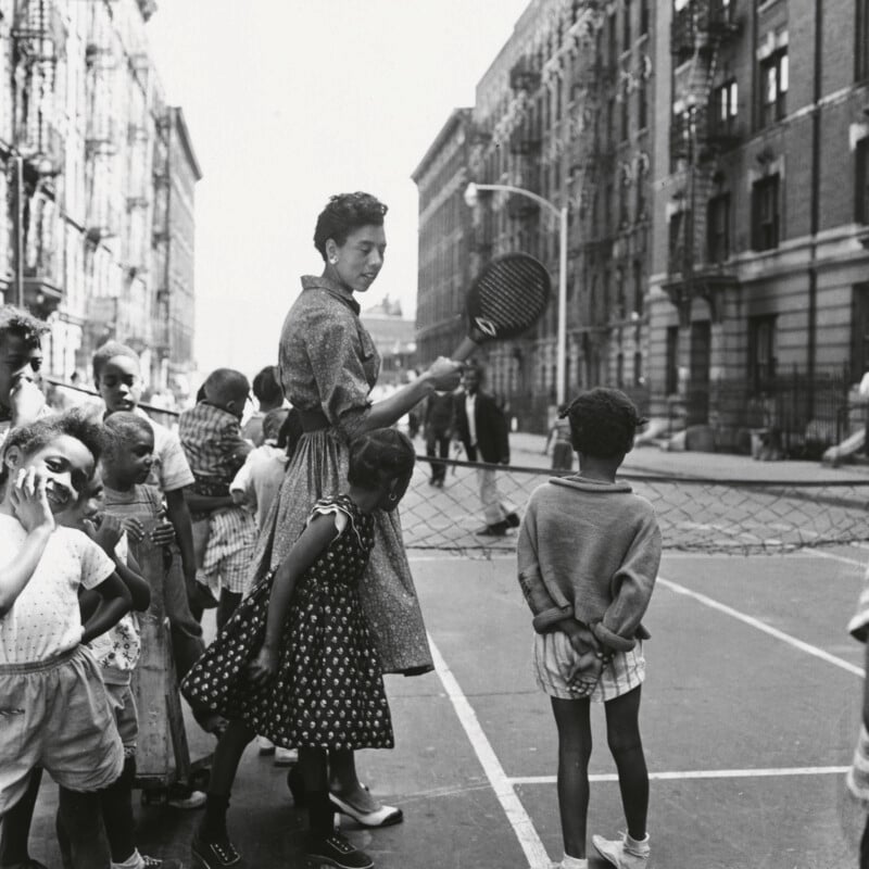 A woman holding a tennis racket stands on a city street court surrounded by a group of children, with apartment buildings in the background.