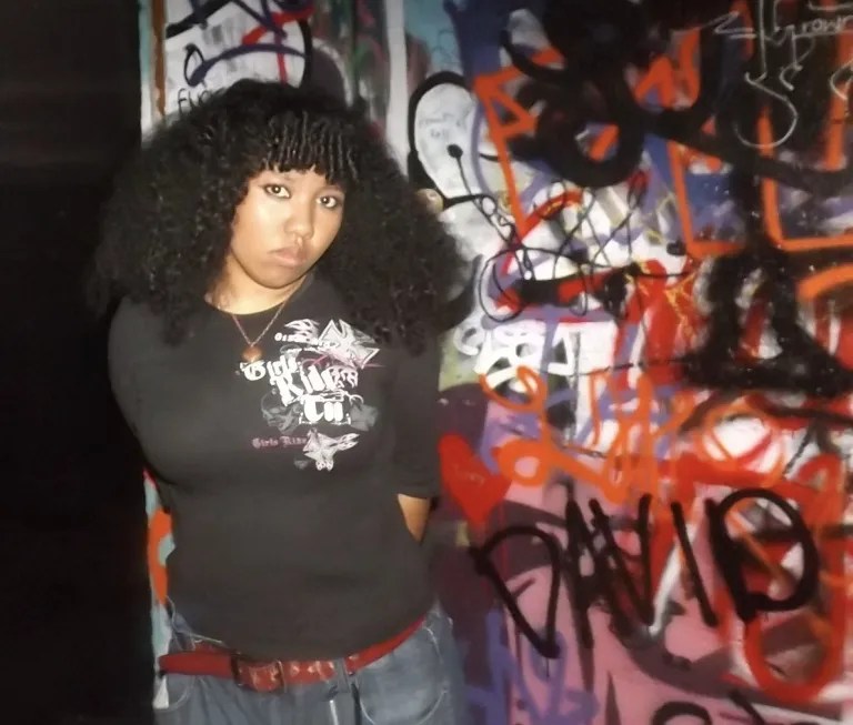 A woman with voluminous curly hair stands against a wall covered in colorful graffiti, wearing a graphic T-shirt and jeans; her direct gaze feels calm and self-assured.