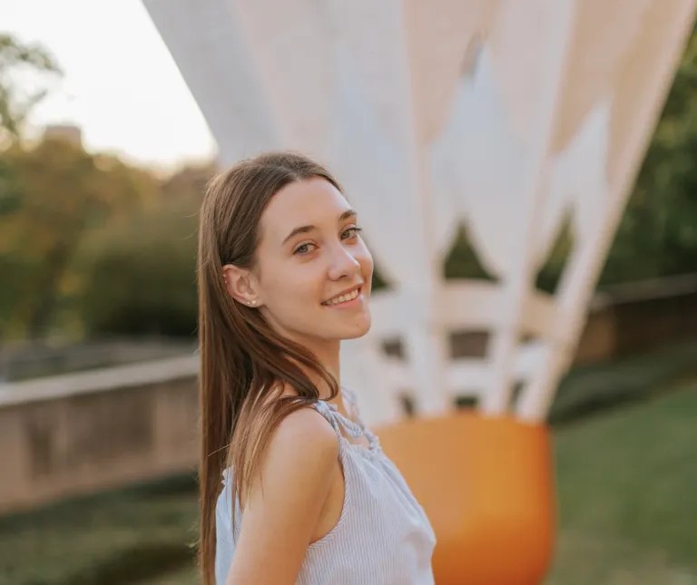 A young woman in a light blue sleeveless dress smiles over her shoulder on a white modern bridge, with greenery behind her in warm evening light.