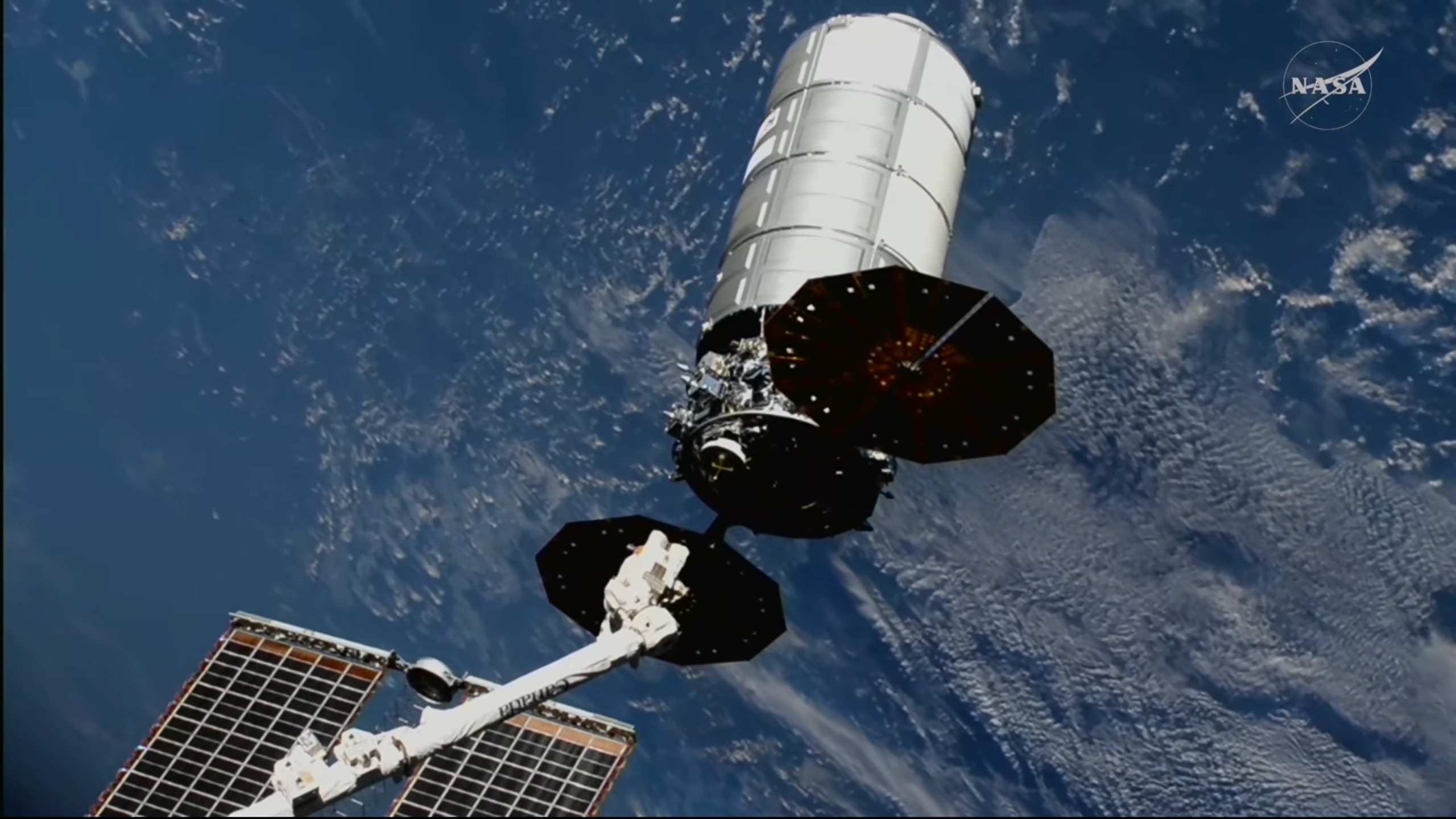 a skinny white robotic arm releases a large cylindrical silver spacecraft in orbit, with earth in the background