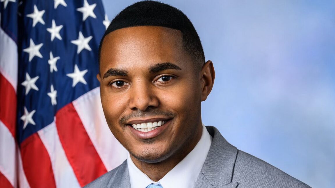 A man smiles in a suit in front of an American flag