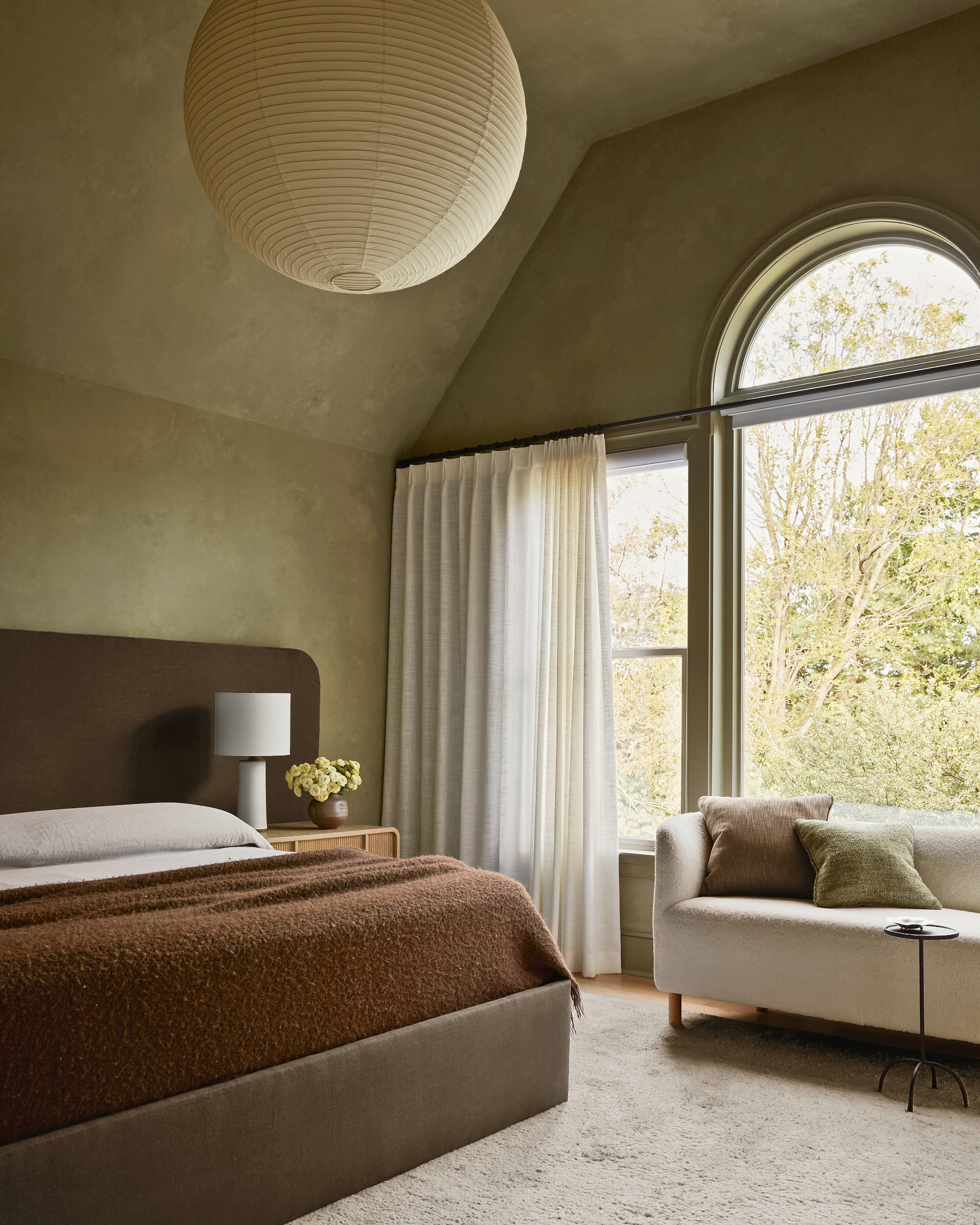 Bedroom with green plaster walls, dark brown oversized headboard, russet throw, cream curtains and cream sofa with small black side table