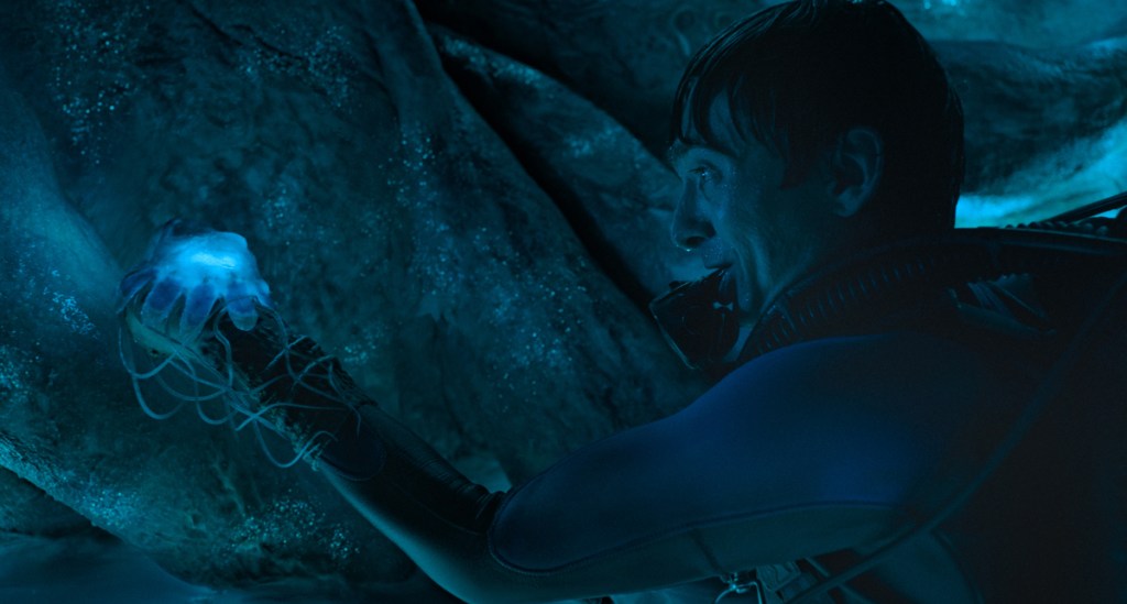 A diver in a dark cavernous area holds a glowing sea creature on an extended arm
