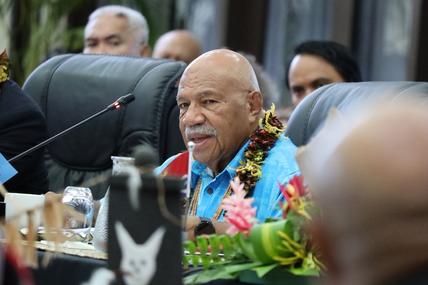 A man wearing a bright blue island shirt with a garland around his neck sits in front of a microphone.