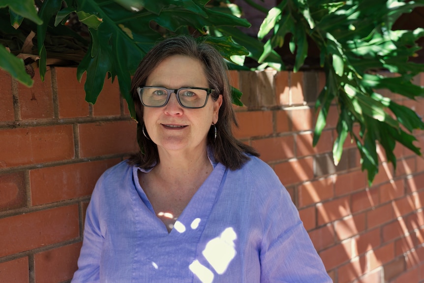 A woman with dark hair and dark framed glasses in a light blue shirt stood outside near red brick wall