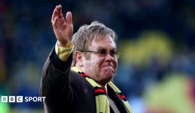 Elton John wearing a yellow scarf with red and black stripes, waving on the pitch at Vicarage Road