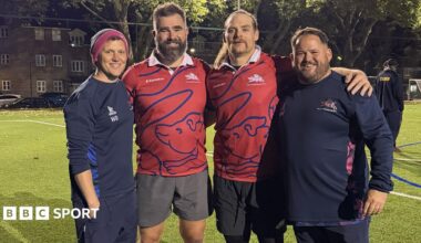 Jason Kelce and Beau Allen with arms round each other and two members of Old Streetonians RFC staff on training pitch under floodlights. Both NFL players are wearing red playing tops.