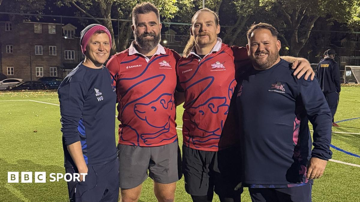 Jason Kelce and Beau Allen with arms round each other and two members of Old Streetonians RFC staff on training pitch under floodlights. Both NFL players are wearing red playing tops.