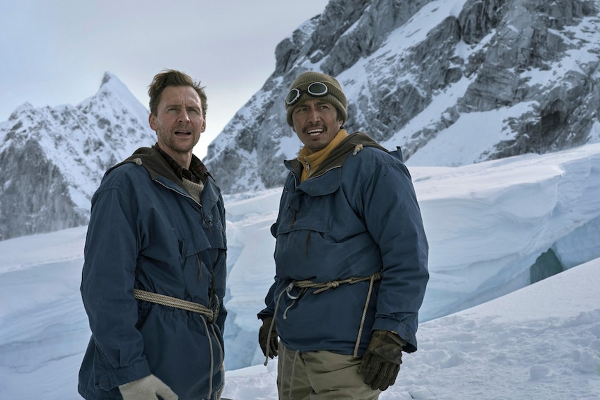 Two men (Tom Hiddleston and Genden Phunstok) stand on a snowy mountainside looking into the distance.