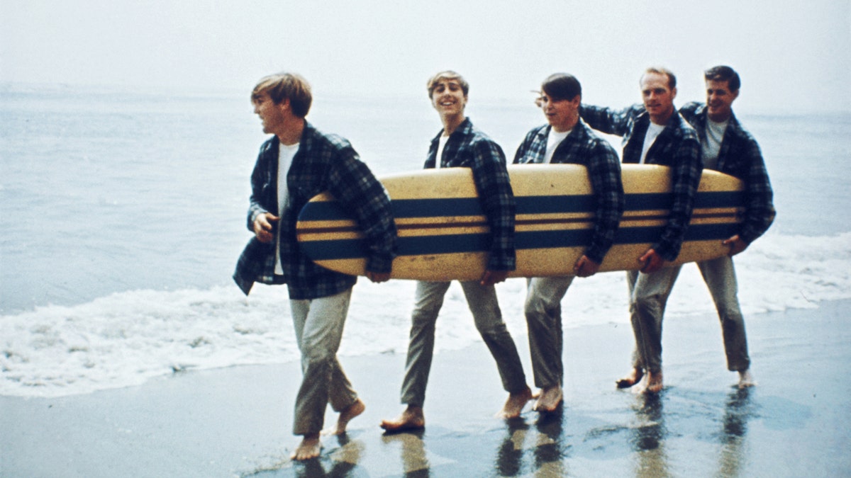 The Beach Boys hold a surf board on a beach