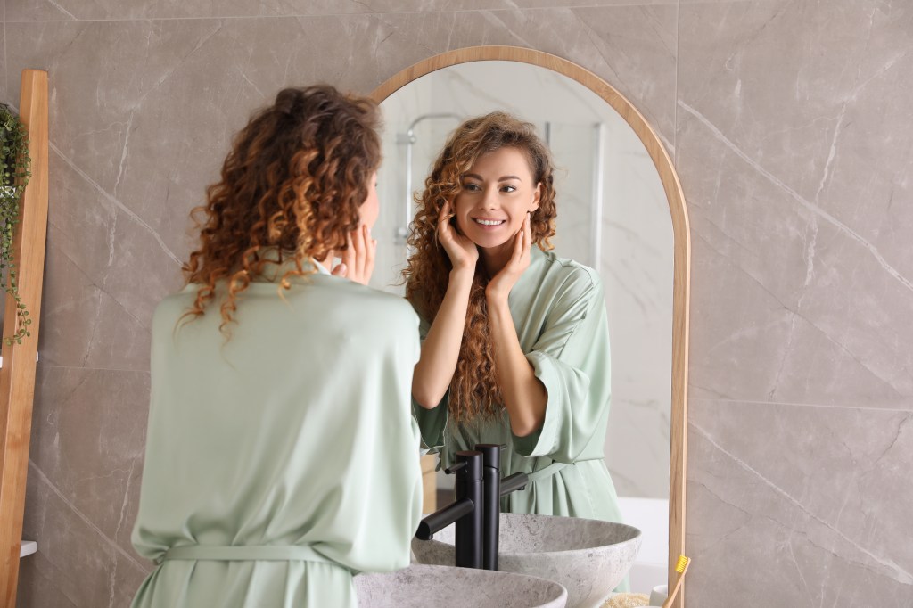 A smiling woman wearing a green robe looks at her reflection in a bathroom mirror, holding her face with her hands.