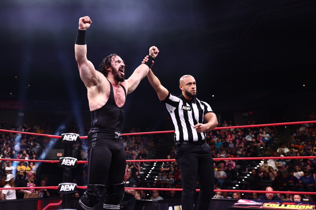 Wrestler PAC celebrates in the ring with an AEW referee holding his arm aloft.