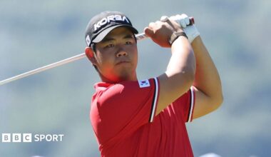 Tom Kim in a red polo shirt and a black cap, both featuring badges with the flag of South Korea. His club is behind his head at the conclusion of his swing