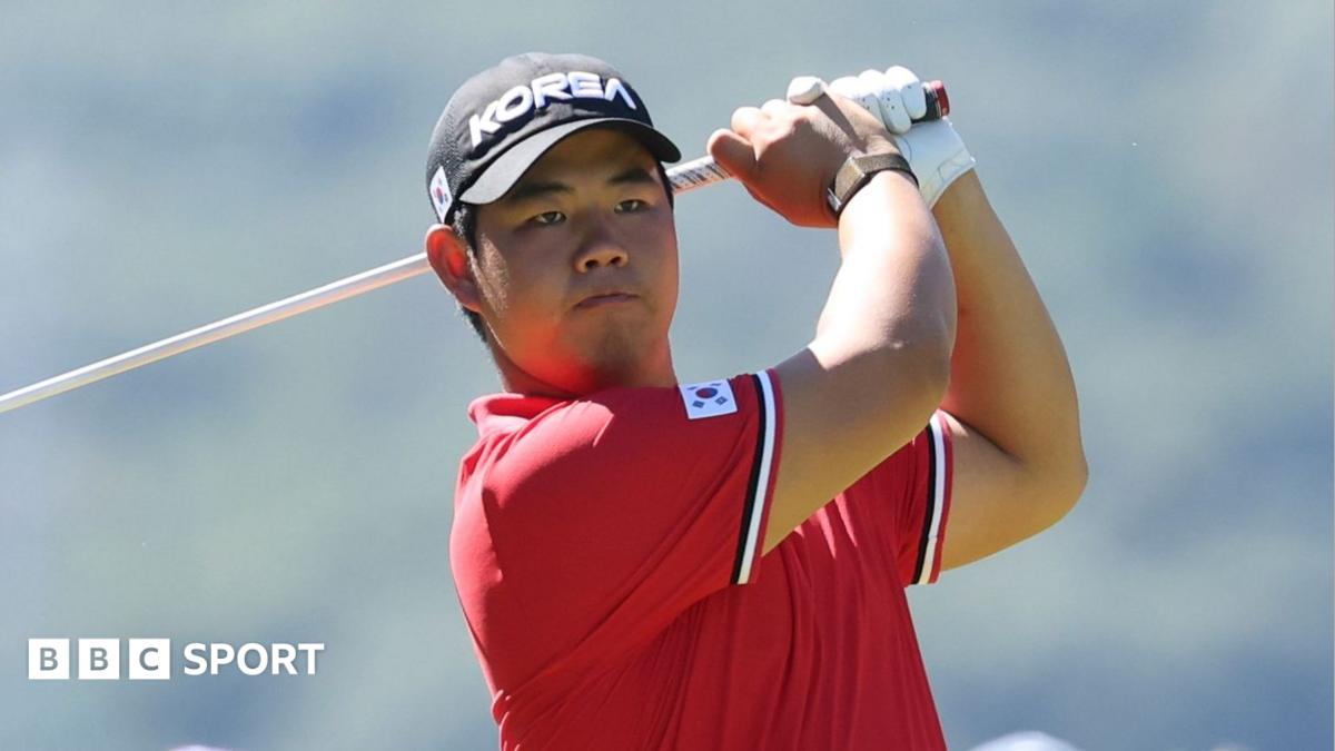 Tom Kim in a red polo shirt and a black cap, both featuring badges with the flag of South Korea. His club is behind his head at the conclusion of his swing